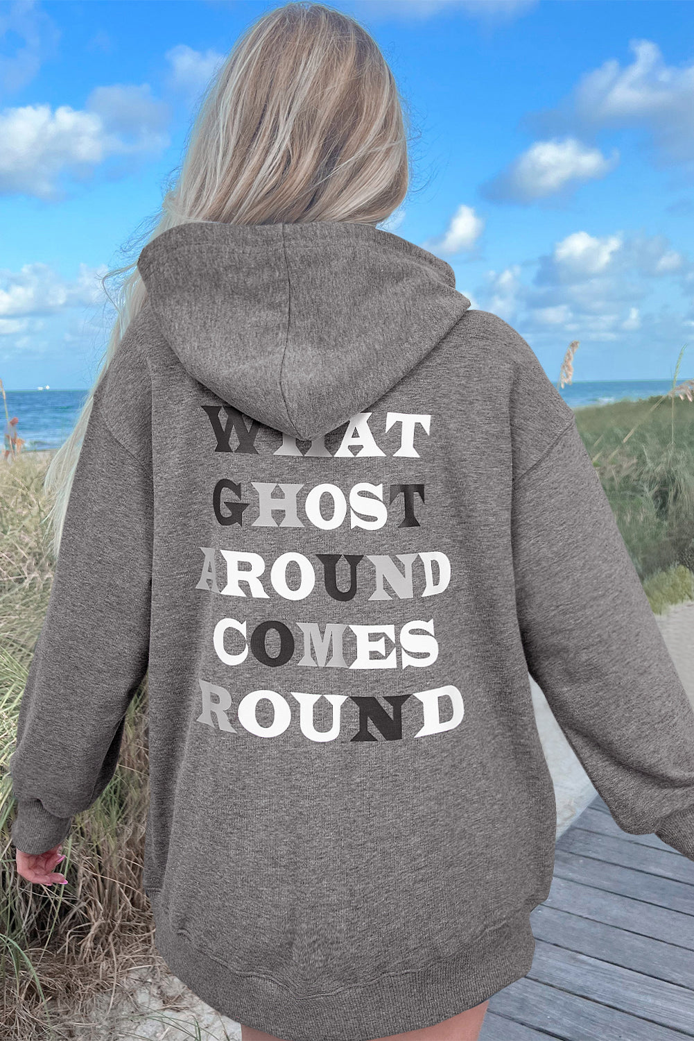 Person wearing a gray hoodie with text on the back, standing by a beach.