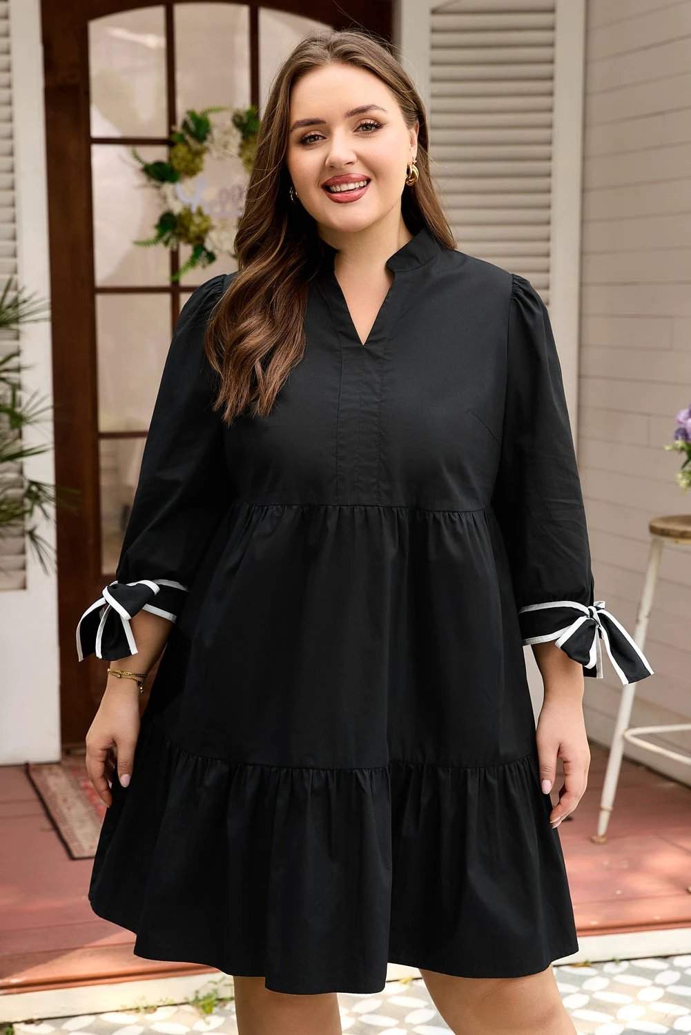 Plus size woman wearing black V-neck mini dress with contrast trim and knot cuffs standing against white background
